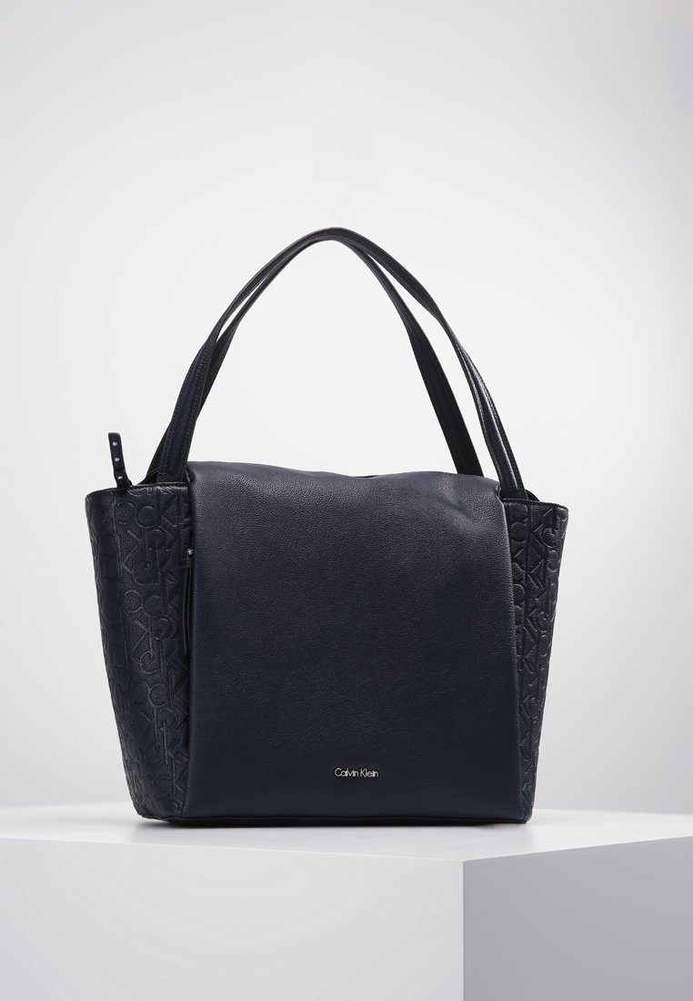 Sac à main en cuir noir Calvin Klein avec des côtés en logo embossé et des poignées doubles, posé sur un piédestal blanc contre un fond gris.