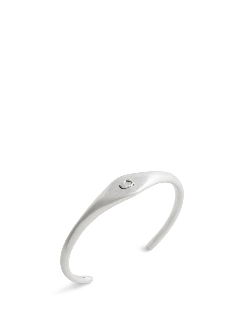 Bracciale a manica in metallo argento con un design liscio e curvo; presenta un piccolo accento circolare vicino alla parte superiore e una finitura testurizzata.