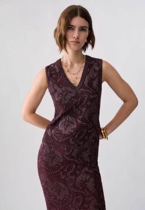 Giovane donna con capelli corti castani che indossa un vestito di maglia viola floreale senza maniche, collane dorate a strati e braccialetti dorati a un polso.