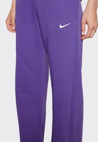 Lila sweatpants med avslappnad passform, tillverkade av mjukt tyg. Har en vit Nike-logotyp på vänster lår. Inga synliga mönster eller detaljer.