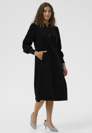 Schwarzes Cordkleid mit Kragen, Knöpfen und Seitentaschen. Lange Ärmel und knielanger Saum. Kombiniert mit hellgrauen spitzen Absätzen.