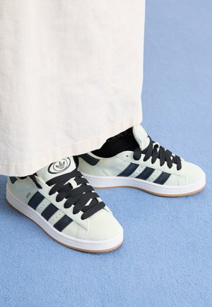 Białe sneakersy Adidas z czarnymi paskami i sznurowadłami, noszone z czarnymi skarpetkami, stojące na niebieskim dywanie, widoczny dół białych spodni.