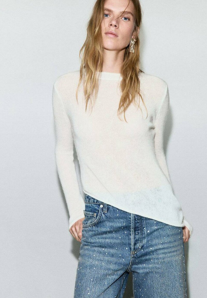 Suéter blanco ligero y translúcido de mangas largas, combinado con jeans azules que tienen pequeñas motas blancas y un ajuste ligeramente relajado.