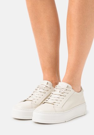 Vista a metà polpaccio di una persona che indossa sneakers bianche con plateau e lacci, con tomaia in pelle liscia su sfondo bianco.