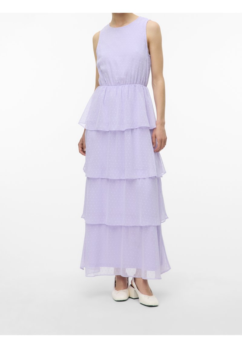 Robe maxi lavande sans manches à étages avec un motif discret à pois, portée avec des chaussures blanches à talons carrés sur fond blanc.