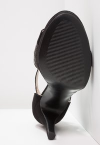 Černá botka na vysokém podpatku s texturovaným semišovým svrškem a elegantním, zakřiveným podpatkem. Podešev má hladký povrch a vertikální drážky pro lepší přilnavost.