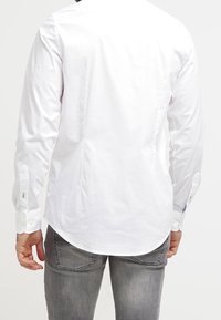 Witte lange mouwen blouse met een afgeronde zoom, voorzien van manchetknopen en een gladde textuur. Draagt grijze spijkerbroek, gezien van achteren.