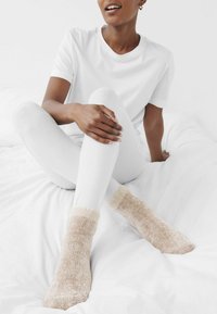 Gemütliche, hellbeige Fuzzy-Socken aus weichem, strukturiertem Stoff mit einem flauschigen Bund, kombiniert mit weißen gerippten Leggings und einem schlichten weißen T-Shirt.