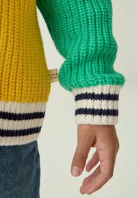 Strickpullover mit gelben und grünen Einsätzen, ausgestattet mit gerippten Bündchen in Marineblau und Weiß. Etikett an der Seitennaht sichtbar.