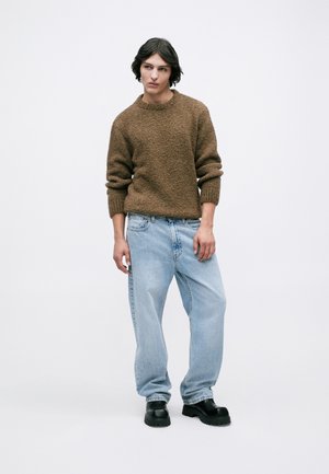 Jovem vestindo um suéter texturizado castanho, jeans largos azul-claro e botas chunky pretas, em pé contra um fundo branco liso.