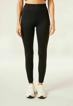 Leggings neri realizzati in tessuto elastico, design a vita alta, texture liscia, silhouette aderente, abbinati a scarpe da ginnastica bianche.
