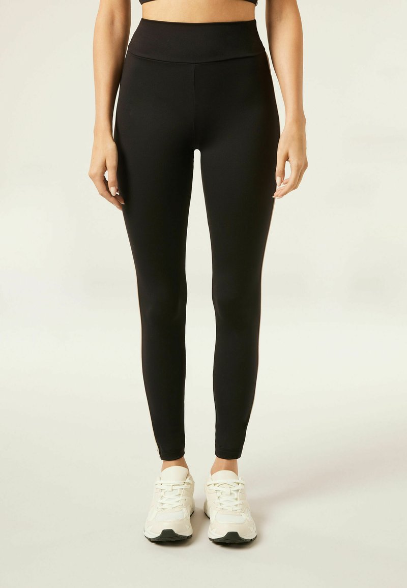 Leggings noirs en tissu extensible, taille haute, texture lisse, silhouette moulante, associés à des chaussures de sport blanches.