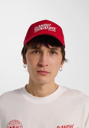 Jong persoon draagt een rode pet met de tekst "Danish Dynamite", wit shirt met rood logo en grote oorbellen, tegen een effen lichte achtergrond.
