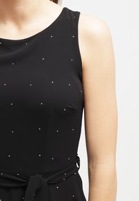 Musta hihaton mekko, jossa on vartaloa myötäilevä yläosa, koristeltu pienillä hopeisilla pisteillä ja solmittu vyötärö yksivärisessä, sileässä kankaassa.