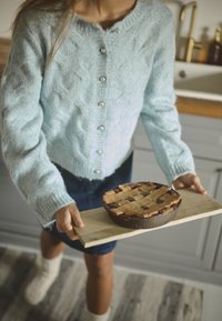 Cardigan azzurro chiaro a maglia con bottoni perlacei, che tiene una torta con crostata a reticolo su un tagliere di legno. La torta ha una crosta e un ripieno di colore marrone scuro.
