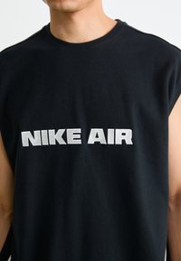 Moški nosi brezrokavnik črne barve z velikim belim napisom "NIKE AIR" na sredini prsnega predela, v ozadju pa je enobarvna bela podlaga.