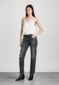 Hvit camisole med blonde detaljer ved halsen, kombinert med svarte jeans. Modellen har på seg svarte ankelstøvler og briller. Enkelt bakgrunn.