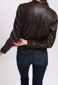 Brązowa skórzana kurtka bomberka z prążkowanym kołnierzem i mankietami. Noszona na ciemnych dżinsach z zakrzywionymi tylnymi kieszeniami, widziana od tyłu.