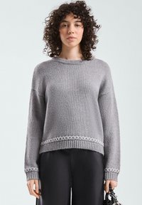 Grauer Strickpullover mit rundem Ausschnitt, überschnittenen Schultern und gerippten Bündchen. Verziert mit einem dekorativen weißen Rautenmuster entlang des Saums.