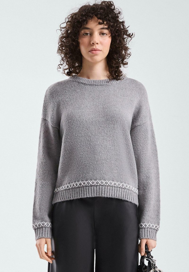 Grauer Strickpullover mit rundem Ausschnitt, überschnittenen Schultern und gerippten Bündchen. Verziert mit einem dekorativen weißen Rautenmuster entlang des Saums.