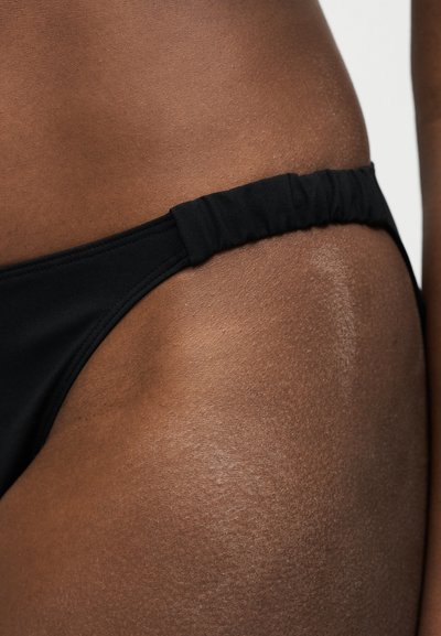 Κοντινό πλάνο του ισχίου ενός ατόμου που φοράει μαύρο κάτω μέρος μπικίνι με ραβδώσεις ελαστικών πλαϊνών λουριών σε σκούρο καφέ δέρμα.