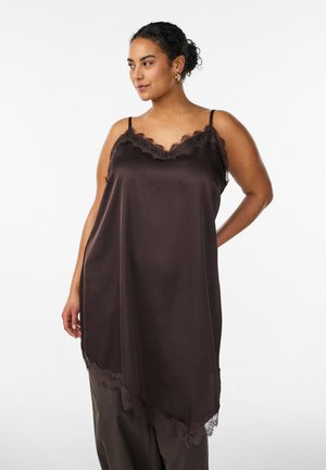 Žena v voľných tmavohnedých saténových šatách s čipkovaným lemom, tenkými ramienkami a tmavých nohaviciach, pózujúca pred jednoliatym pozadím.