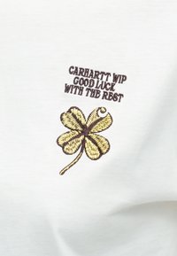 Biele tričko s vyšitým štvorlístkom v žltej a hnedej farbe, spolu s textom "CARHARTT WIP VEĽA ŠŤASTIA DO ZBYTOKU."
