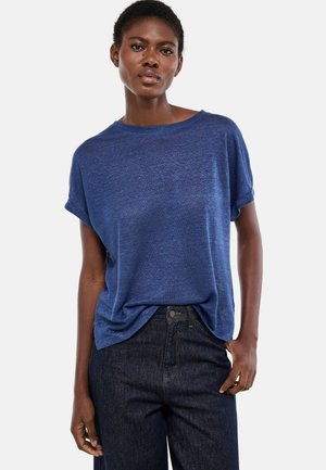 Femme portant une chemise bleue à manches courtes ample, légèrement rentrée dans un jean foncé taille haute, debout devant un fond uni.