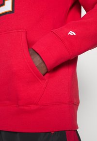 Main à peau foncée dans la poche d'un sweat-shirt rouge vif avec un petit logo blanc sur le poignet de la manche.
