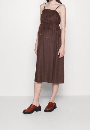 Kvinde iført en ærmeløs brun midi-kjole med tynde stropper og bindebånd i taljen, kombineret med brune hælede træsko mod en ensfarvet baggrund.