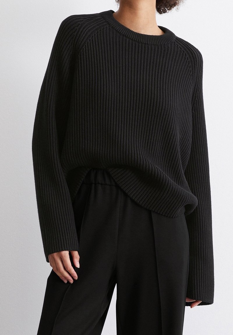 Personne portant un pull en maille côtelée noire à manches longues et un pantalon noir ample sur un fond clair uni.