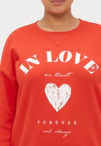 Raudona džemperis su baltu tekstu, kuriame parašyta "MEILĖJE tikime", virš širdies grafikos, su "AMŽINAI ir visada" apačioje mažesniu šriftu.