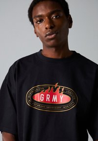 Camiseta de algodón negra con un gran gráfico de llamas y el texto "GRMY" en el centro. Mangas cortas, corte holgado, cuello redondo.