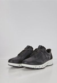 Svarte atletiske sko laget av syntetisk materiale, med en teksturert overdel, kontrasterende hvit mellomsåle og en lavprofildesign med snøring.