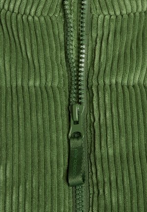 Tessuto in velluto a coste verde con una cerniera in plastica parzialmente aperta, caratterizzata da una linguetta in tessuto verde con la scritta "new balance".