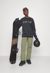 Snowboard avec une base noire, un graphisme sombre et des fixations rouges. Le modèle porte un sweat-shirt noir avec "BRTN", un pantalon olive, des gants bleus et un casque noir.