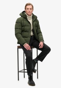 Giacca a vento verde con cappuccio, polsini a costine e bottoni neri, abbinata a un dolcevita beige, pantaloni neri e stivali neri con lacci.