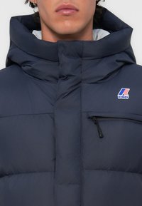 Marineblauwe pufferjack met een hoge kraag, een ritszak op de borst en het K-Way-logo. Kenmerkt zich door een zachte, gewatteerde textuur met een glanzende afwerking.