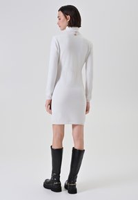 Donna con capelli corti e scuri, vestita con un abito bianco a maniche lunghe e collo alto, e stivali neri fino al ginocchio, in piedi di spalle.