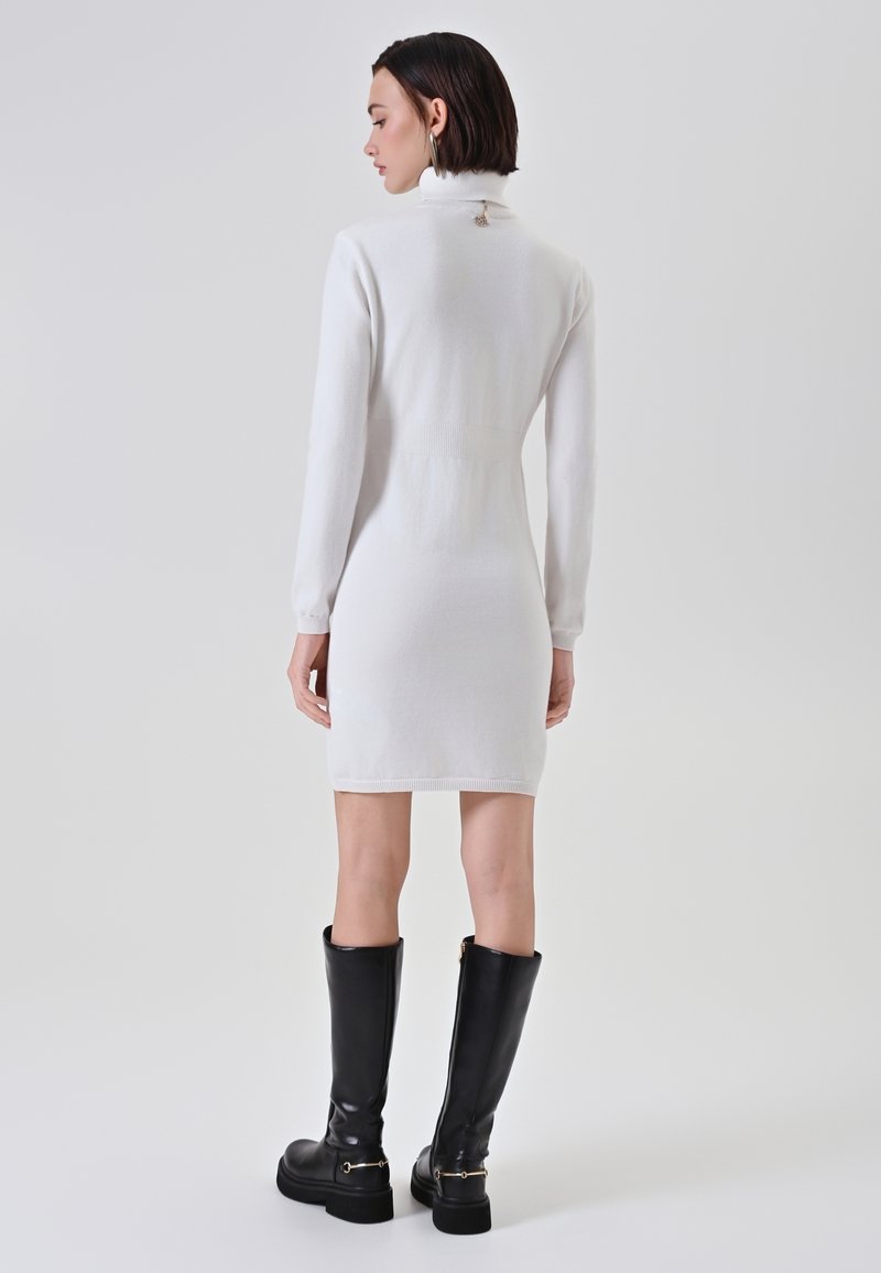 Donna con capelli corti e scuri, vestita con un abito bianco a maniche lunghe e collo alto, e stivali neri fino al ginocchio, in piedi di spalle.