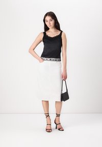 Črna satenasta top z čipkastim robom v kombinaciji z belo krilom do gležnjev; črne sandale na vezanje in majhna črna torbica dopolnjujeta obleko.