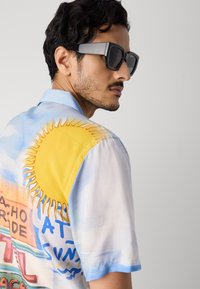 Majica s kratkimi rokavi v pastelno modri barvi s sončnim grafikom in pisanimi napisi. Ima sproščen kroj, gladko teksturo in kontrastni ovratnik.