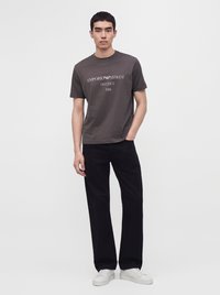 Млад мъж стои с една ръка в джоба, носейки тъмна тениска Emporio Armani, черен панталон и бели маратонки на едноцветен фон.