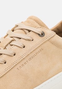 Beige suede sneaker with textured surface, matching laces, and embossed logo on the side. White rubber sole and minimalistic design.