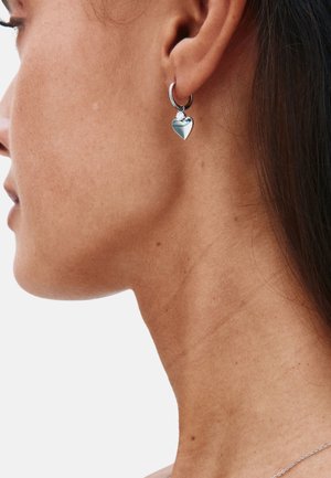 Orecchino a cerchio in argento con un piccolo pendente a forma di cuore, abbinato a un dettaglio di cristallo, visto da vicino di lato su uno sfondo semplice.