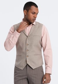 Chaleco de mezcla de lana beige con un ajuste entallado, que cuenta con cinco botones, dos bolsillos frontales y un dobladillo puntiagudo, llevado sobre una camisa rosa claro.