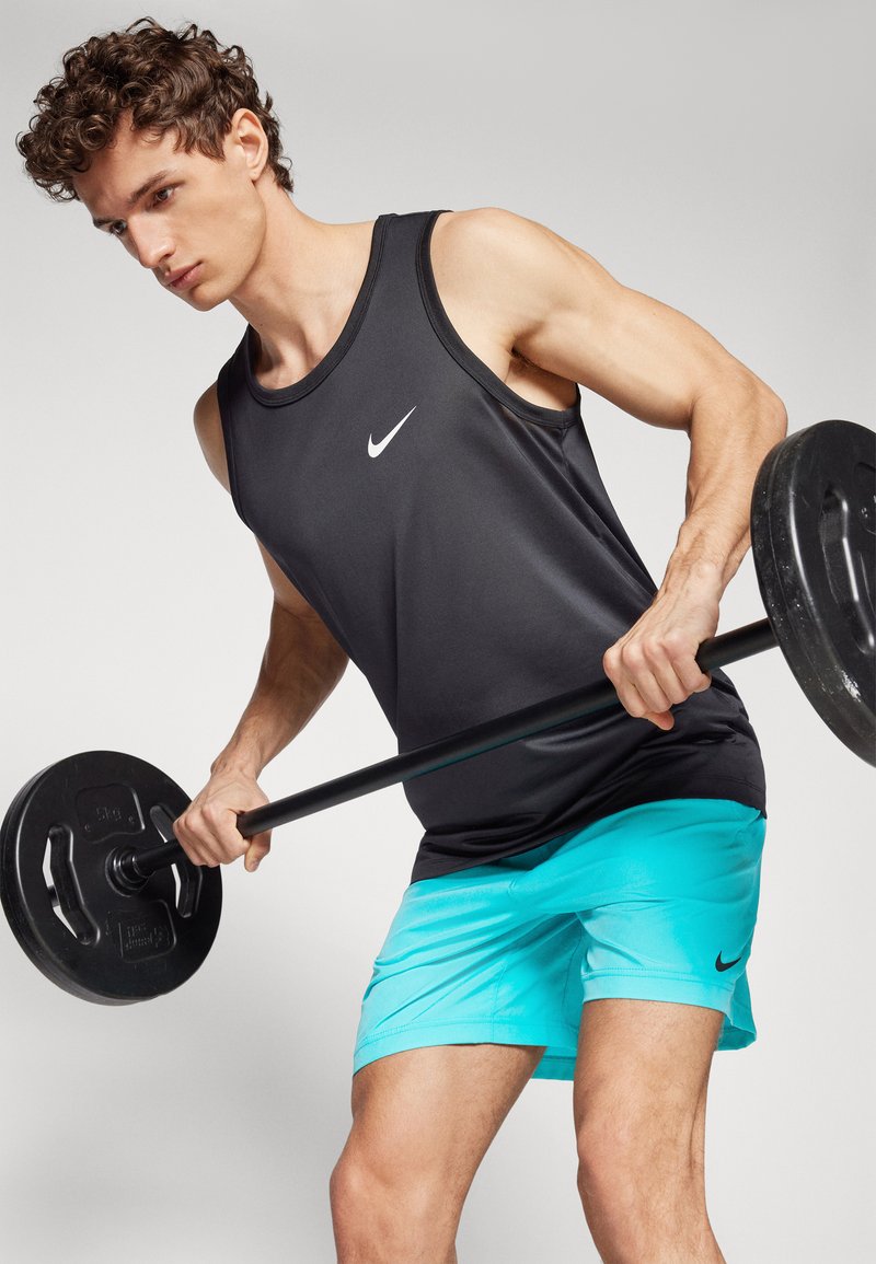 Top atlético negro sin mangas con un logo blanco de Nike, combinado con pantalones cortos turquesa. El modelo está levantando una barra con pesas negras.