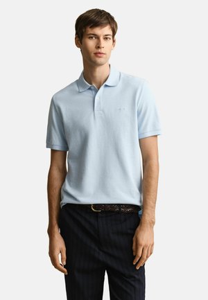 Jonge man die een lichtblauw poloshirt met korte mouwen draagt, ingestopt in een donkergekleurde broek met krijtstrepen en een bruine gevlochten riem, staand tegen een witte achtergrond.