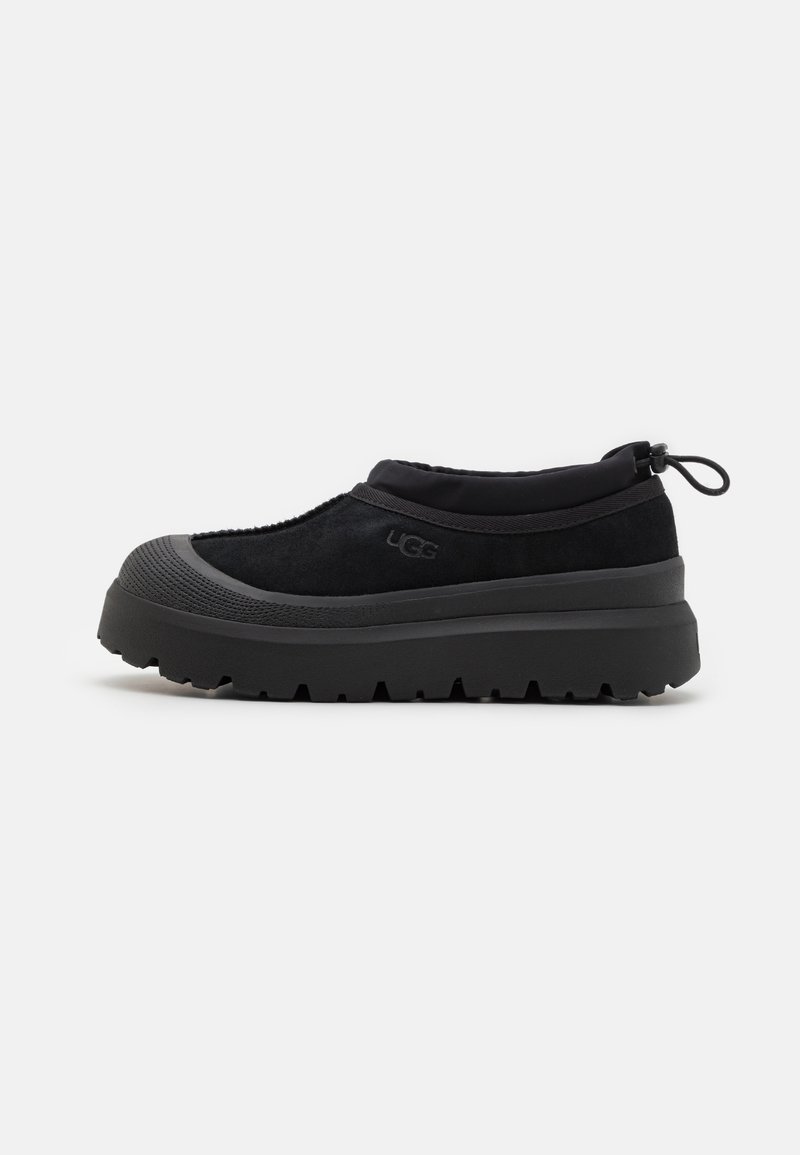 Black slip-on shoe featuring a suede upper, rubber sole with a rugged tread pattern, and a pull tab at the back for easy wear.