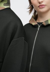 Fekete kapucnis pulóver cipzárral az elején, terepmintás szegéllyel rendelkező kapucnival és túlméretezett szabással, puha, texturált anyagból készült.